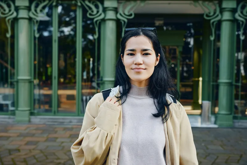 Woman in casual outfit outdoors near a building, wearing a coat and sweater, posing in a calm street setting with soft natural light and urban backdrop
