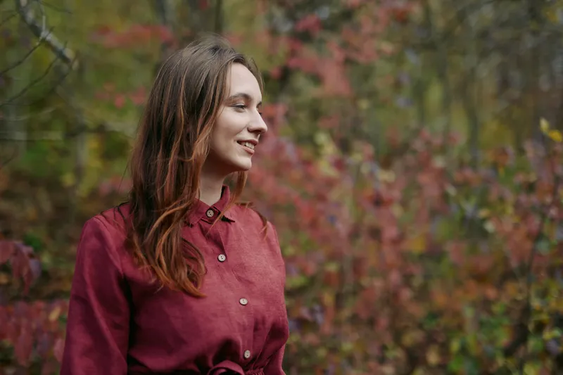 A young woman with a natural smile stands among autumn foliage, captured in an authentic moment that conveys warmth, genuine expression and reliable storytelling for lifestyle and portrait imagery.
