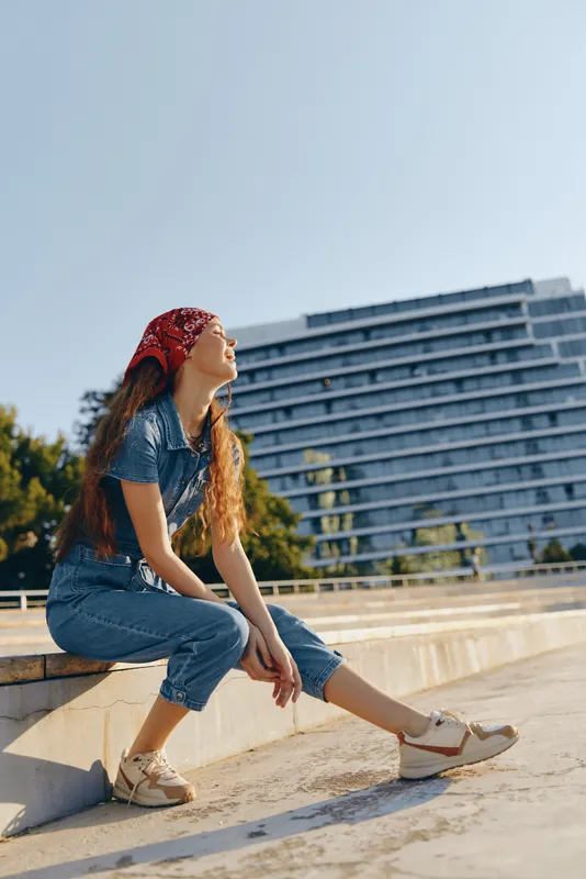 Lifestyle woman in modern boho-western denim street style sits relaxed outdoors, wearing a red bandana and beige sandals under natural sunlight with film color effect.