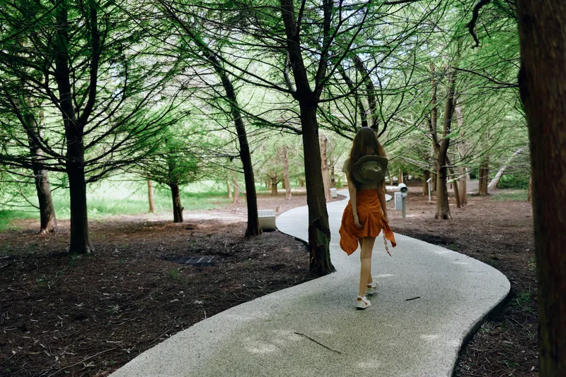 Woman walking on winding path through green forest wearing orange dress and backpack, rear view of a traveler on a curving gravel trail, peaceful nature walk and outdoor exploration.