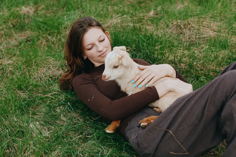 Relaxed woman lying on grass, cuddling a young goat kid outdoors, capturing tender bonding, rural lifestyle, and peaceful self care in soft natural light. It conveys wellness and calm.