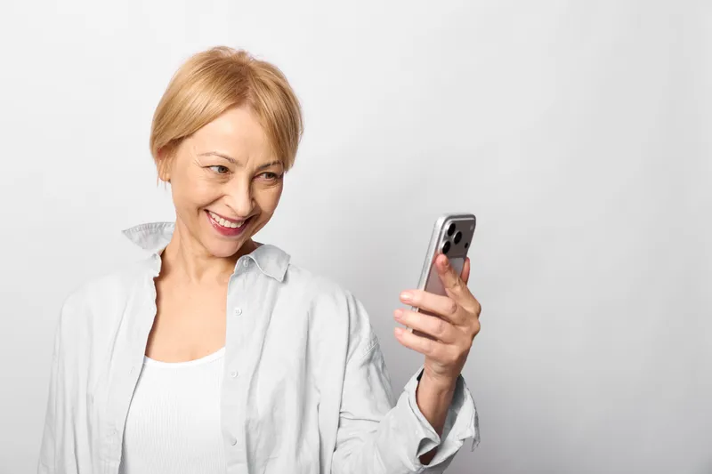 Smiling senior woman holding a smartphone, portrait on plain white background, technology communication with relaxed posture, friendly indoors scene for routine