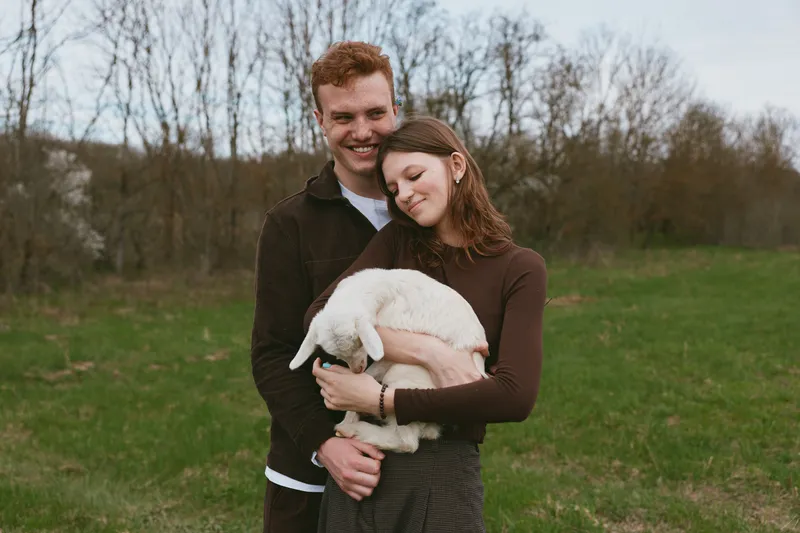 Young couple embraces outdoors while the woman gently holds a white baby goat in a peaceful spring meadow under soft natural light.