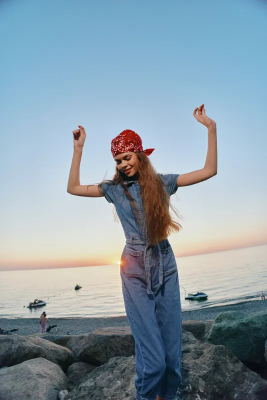 lifestyle woman in modern boho-western denim street style dancing with raised arms at seaside sunset, film color warm tones, casual joyful outdoor moment