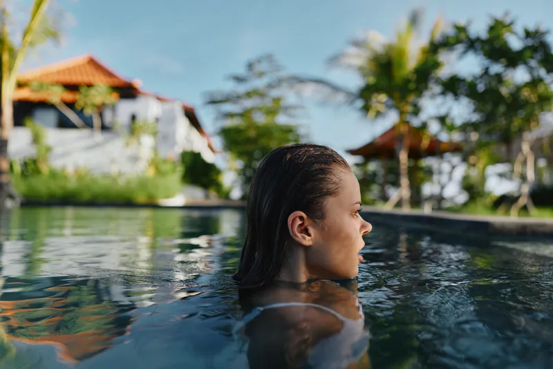 A woman in a pool, side profile, enjoying a sunny tropical resort. Calm water, lush palm trees, and clear sky convey relaxation, leisure, and vacation mood.