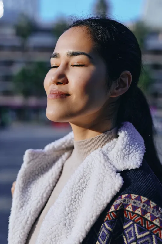 Serene woman basking in sunlight outdoors, wearing a textured coat, enjoying calm relaxation and warmth as daylight illuminates her face with contentment and gentle glow