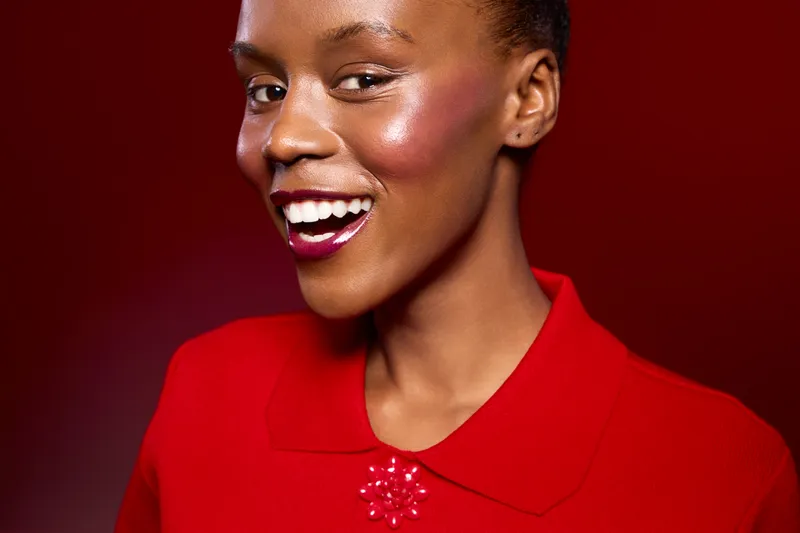 Smiling African American woman with dark skin wearing red blouse with decorative collar on red background. Close-up portrait with bright makeup and joyful expression. Concept of beauty and fashion.