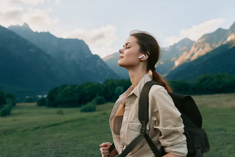 Woman hiker pauses in a scenic meadow with mountains, backpack, listening to music, enjoying fresh air and freedom, relaxed pose, sunny day, nature retreat
