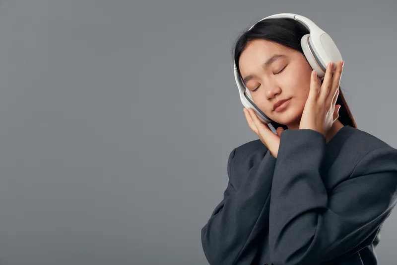 Young woman wearing headphones with eyes closed listening to music, enjoying sound, relaxation and calm in casual dark sweater against gray background.