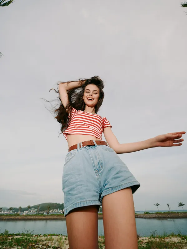 Joyful young woman posing outdoors in a casual summer outfit, striped crop top and high waist denim shorts, smiling brightly as a wide sky and open landscape surround her.