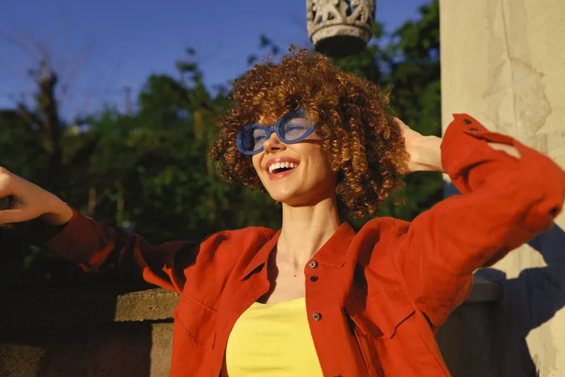 Cheerful woman outdoors in a red jacket, yellow top, and blue sunglasses, smiling under bright sun, relaxed pose, lifestyle moment amid natural greenery and breeze