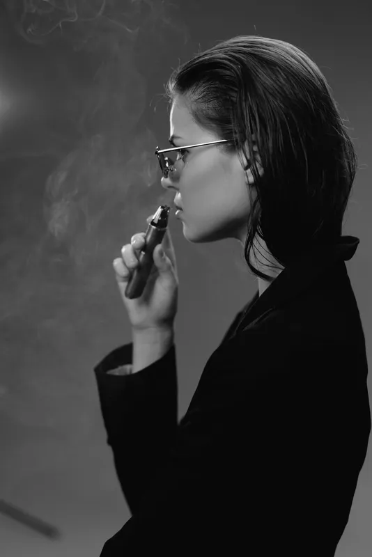 Black and white portrait of a woman vaping, wearing glasses and a dark jacket, with visible smoke and a contemplative mood.