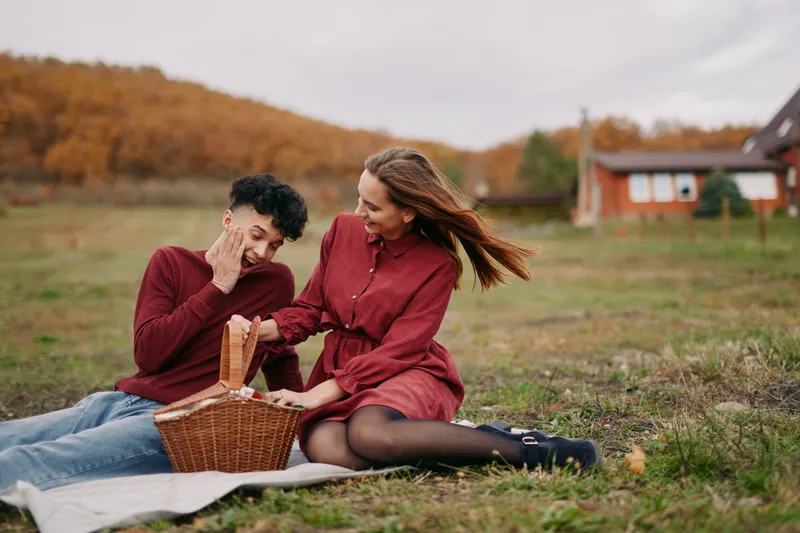 A candid autumn moment captures a couple sharing a picnic on a grassy field, with genuine smiles, natural lighting, a woven basket, warm tones, authentic connection in sunlight.