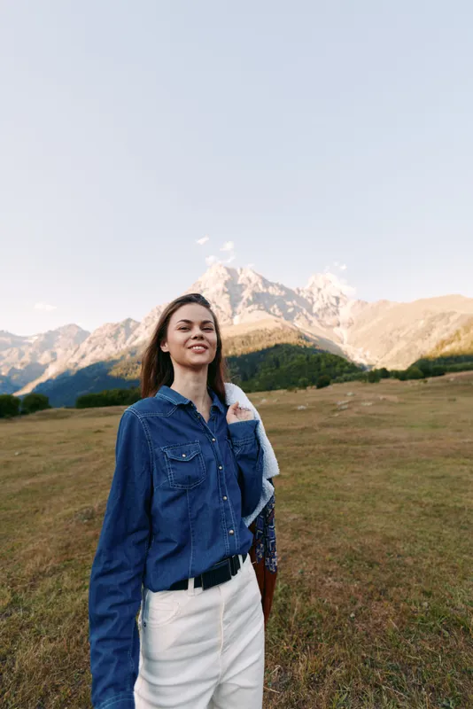 Woman smiling in denim shirt portrait in mountain meadow, casual fashion and travel concept. Outdoor lifestyle, scenic landscape and confident young adult enjoying nature.