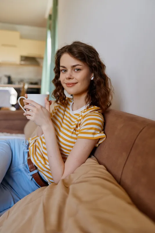Casual woman with curly hair sits on a couch holding a mug in a warm home interior, daytime light. Relaxed lifestyle, comfort, simple self care scene, cozy mood.