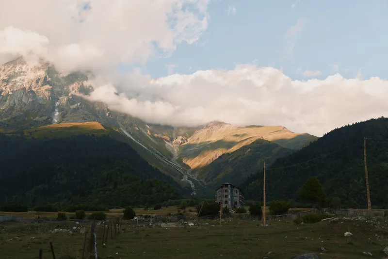 Quiet mountain valley scene shows a small weathered house in a grassy field beneath dramatic clouds with distant peaks forming a rugged backdrop and a sense of solitude.