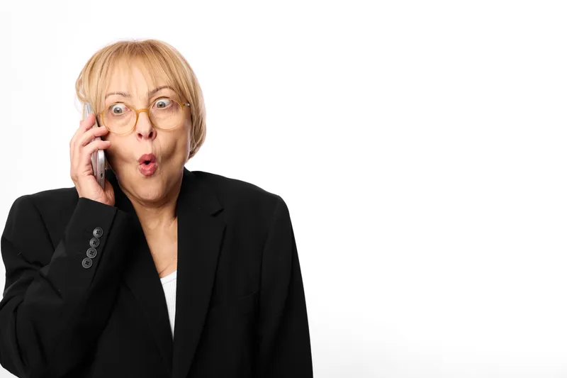 Woman with glasses is surprised while holding a phone to her ear, studio portrait on white background, mature blonde in a business blazer, communication and urgency