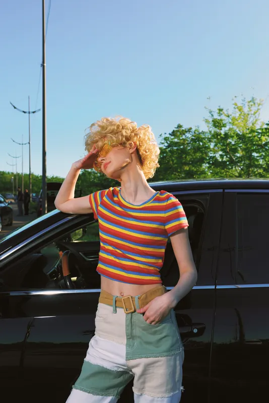 Woman wearing a colorful striped crop top and patchwork shorts stands beside a dark car on a sunny street, exuding retro lifestyle and carefree outdoor fashion