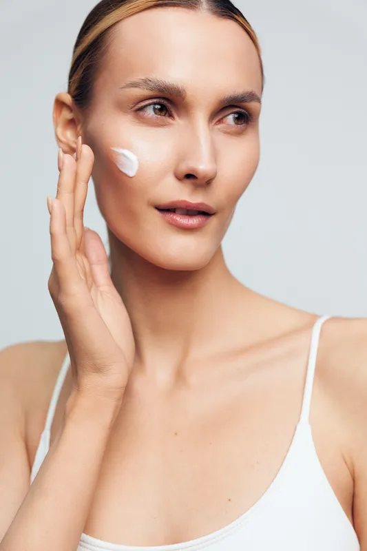 A young woman with clear skin applies white cream to her cheek in a bright studio. The image represents skincare, beauty routine, and natural complexion care.