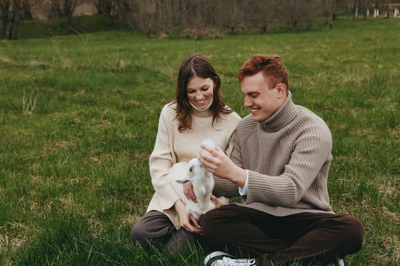 Couple outdoor goat kid affection lifestyle. A smiling pair sit on the grass, gently holding a goat kid amid meadow and trees, wearing cozy sweaters. Warmth, bonding, and nature create a wholesome