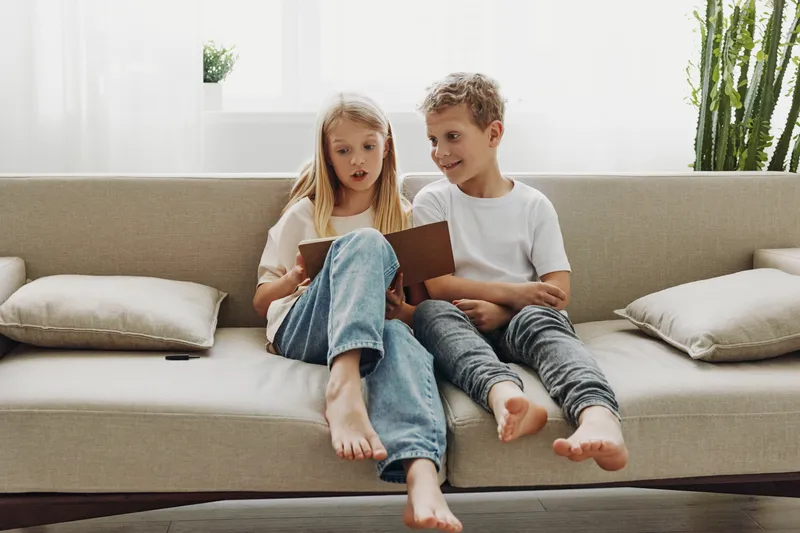Two barefoot children sit on a beige sofa reading a book together in a bright living room, sharing a moment of curiosity and learning on a relaxed weekend.