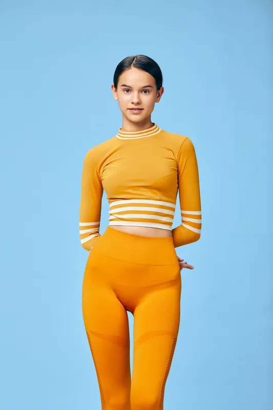 Female fitness model in vibrant orange athletic wear poses against a solid blue background, showcasing sport style, strength and confidence in a contemporary studio.