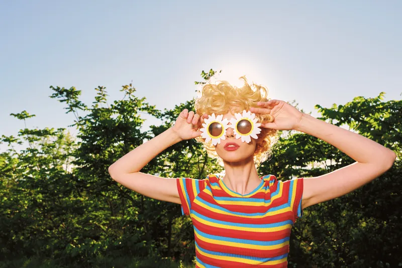 Playful woman outdoors wearing a colorful striped top, holding daisy glasses over her eyes in a sunny park, retro film tones enhancing the carefree mood and vibrant texture.