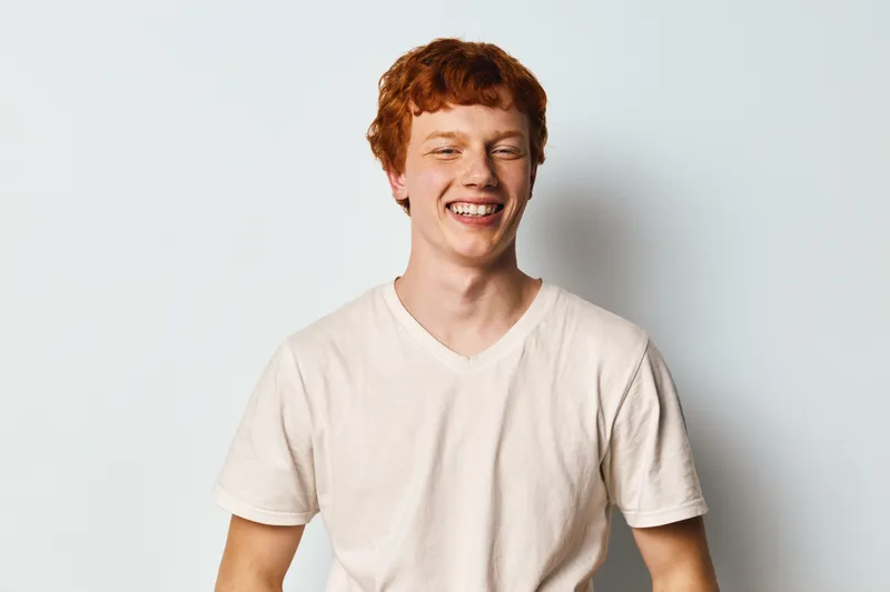 Portrait of red hair young man smiling with closed eyes wearing casual white t shirt on white background, happy emotion expression and relaxed mood in studio light.