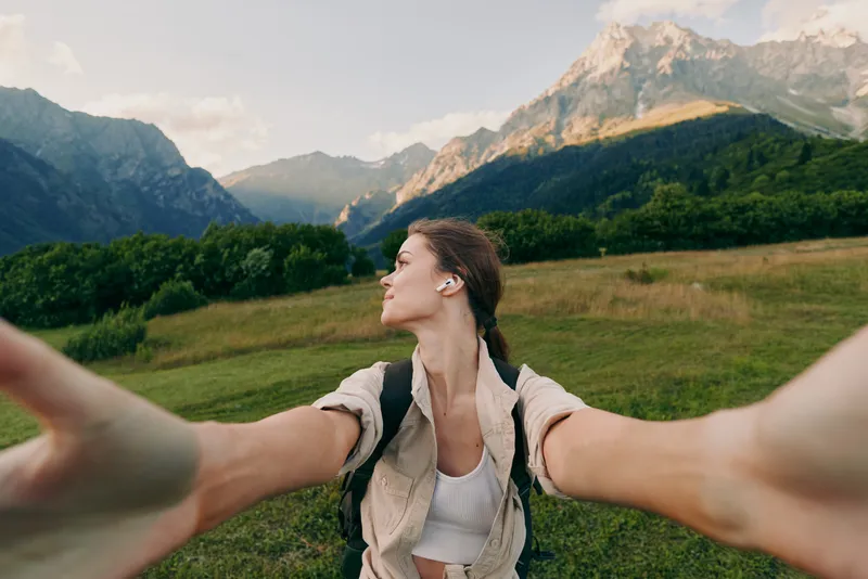 Young woman with backpack takes selfie in mountains enjoys nature fresh air and freedom on sunny day with clear sky and green grass landscape around.