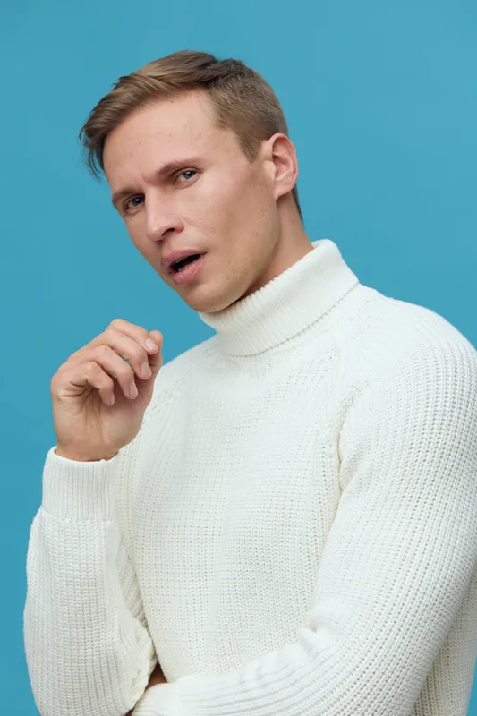 Close-up portrait of surprised man with light brown hair, blue eyes, and fair skin, wearing a white turtleneck sweater, posing against a vibrant blue background, studio shot, casual fashion style. Peo