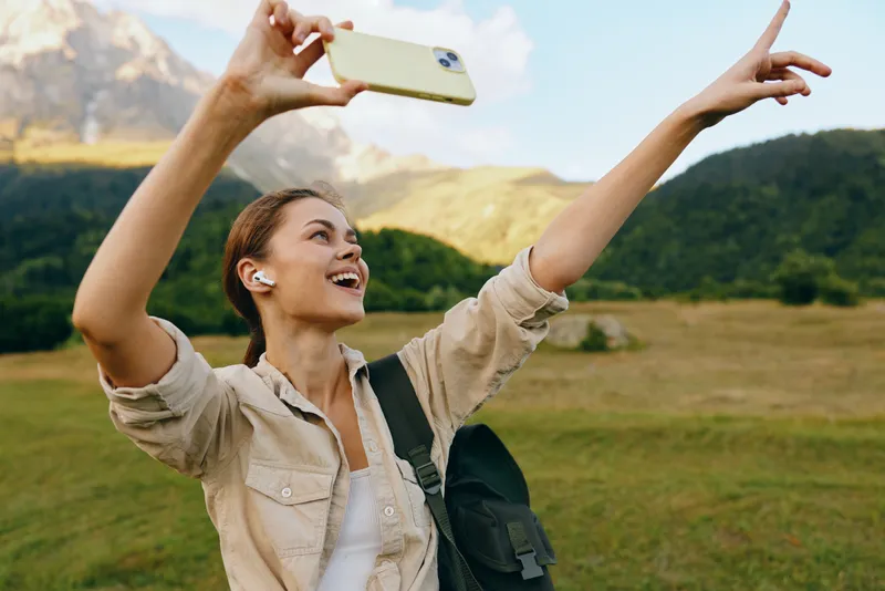 Young woman with short hair takes a selfie with a smartphone while pointing excitedly in a scenic mountain meadow during a sunny day.