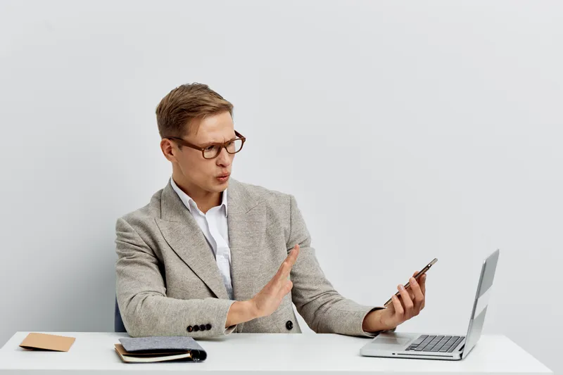 Professional young man in gray blazer and glasses using smartphone at work desk with laptop, notebook, and coffee on plain white background, studio portrait. Business communication concept