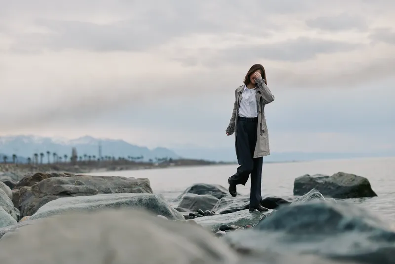 A woman in casual clothing walks on rocks by the sea, covering her face with one hand. The cloudy sky and distant mountains create a calm, contemplative outdoor scene.