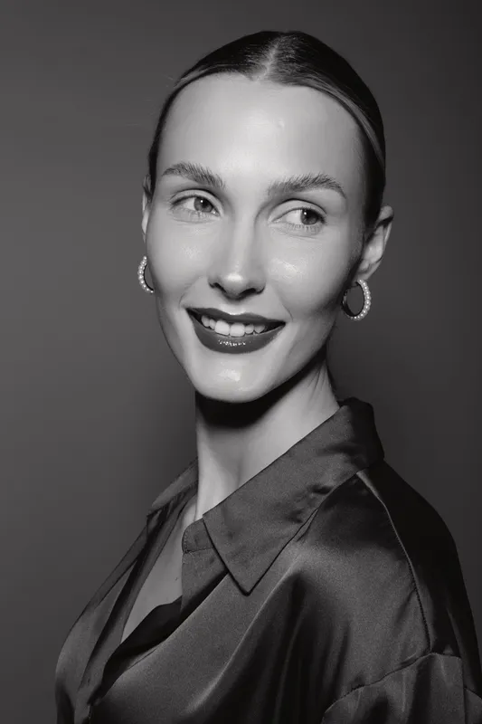 Portrait of a smiling Caucasian woman with fair skin and slicked back hair wearing earrings and a satin blouse. Studio lighting highlights her smooth complexion and elegant style.