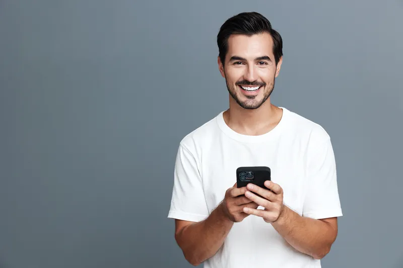 Smiling man holding smartphone, looking at the camera with cheerful expression, wearing casual white t-shirt, isolated on gray background, studio portrait.