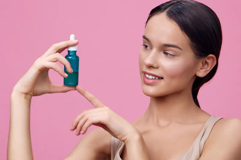 Young woman holding a turquoise serum bottle against a pink background, expressing joy and optimism about skincare and beauty routines