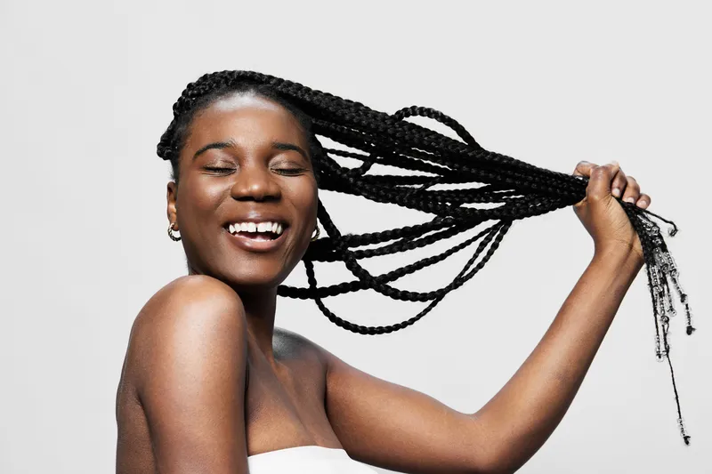 Young Black woman with braids joyfully playing with her hair, showcasing a vibrant smile against a clean, minimalist background Ideal for beauty and haircare concepts