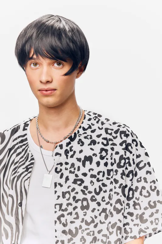 young man with short black hair wearing a stylish patterned shirt and silver chains, looking confidently against a plain white background with soft lighting