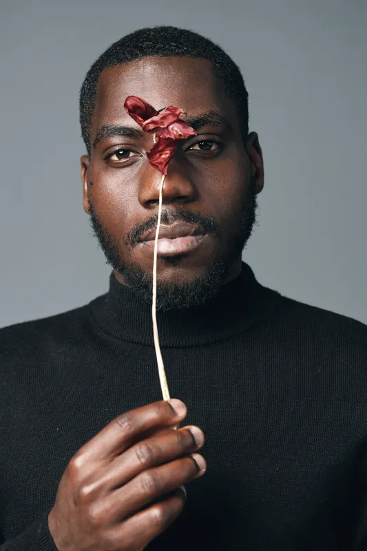 African man with dark skin holding dried red flower in front of face in creative conceptual visual style. Portrait with black turtleneck and neutral background for artistic expression.