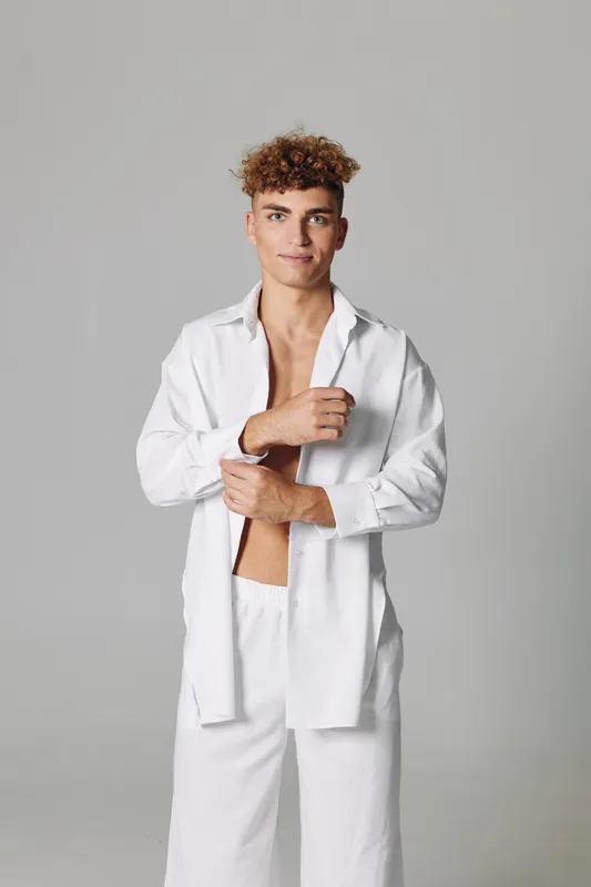 Young man with curly hair wearing white open shirt and pants standing against gray background, looking at camera with confident expression and relaxed posture.