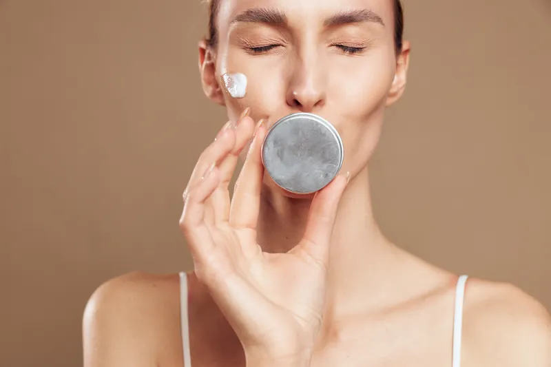 Young woman applying face cream in a neutral studio, close up on a beauty jar and a calm mood, showcasing a skincare routine and self care in soft lighting.
