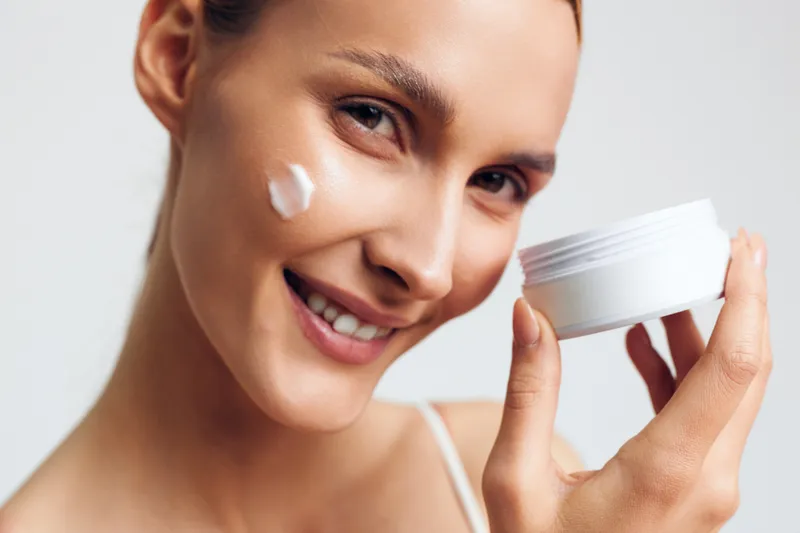 Young woman with clean skin smiling applies face cream on cheek holding white jar close to face isolated on white background beauty skincare concept.