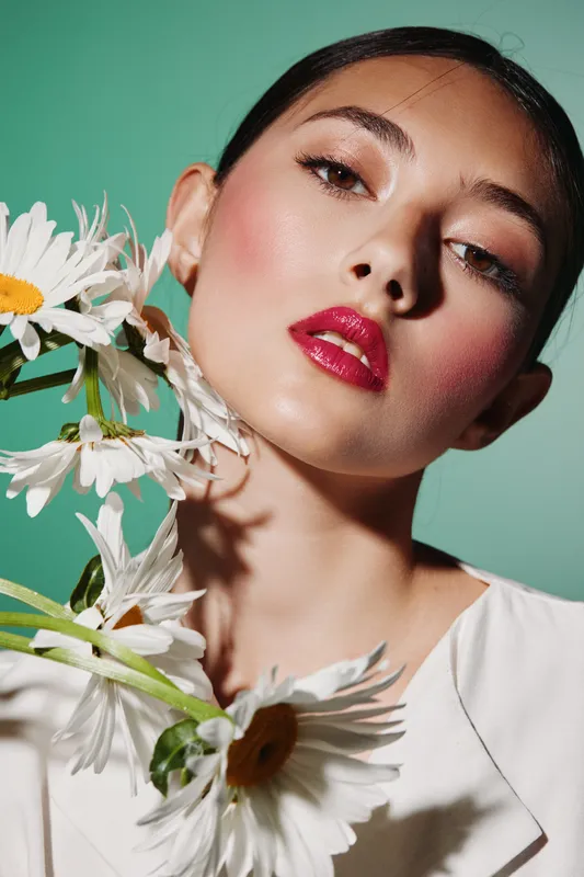Close-up portrait of a fashion model with glossy lips and subtle eye makeup, posing with white daisies against a mint green background. Concept for beauty, fashion, and creative style.