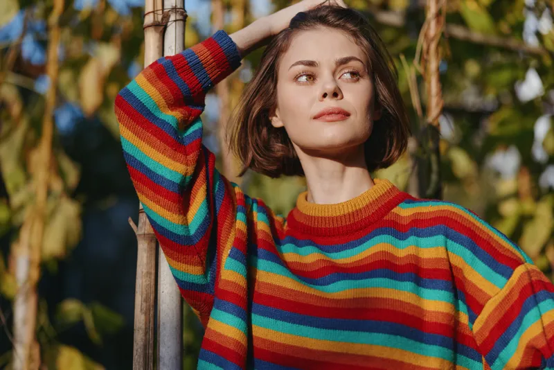 Woman in colorful striped sweater, short brown hair, outdoor portrait of a young adult in casual outfit leaning on bamboo stake, looking thoughtful and softly smiling in garden sunlight