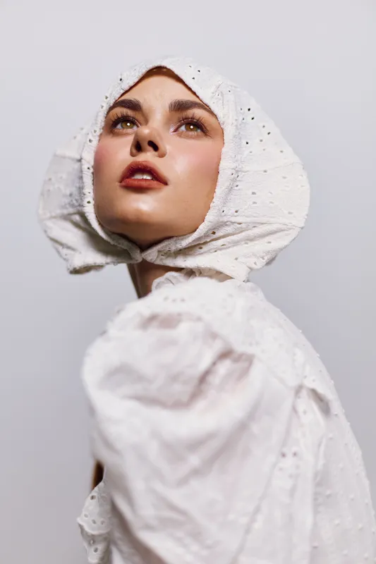 Young woman in a white vintage dress and bonnet, expressing hope and elegance against a minimalist backdrop, perfect for fashion and portrait themes
