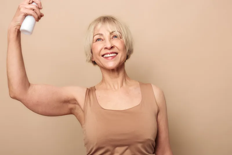 Smiling mature woman with short blonde hair sprays deodorant on her underarm wearing beige tank top. Studio portrait on beige background. Concept of personal care, hygiene, and confidence.