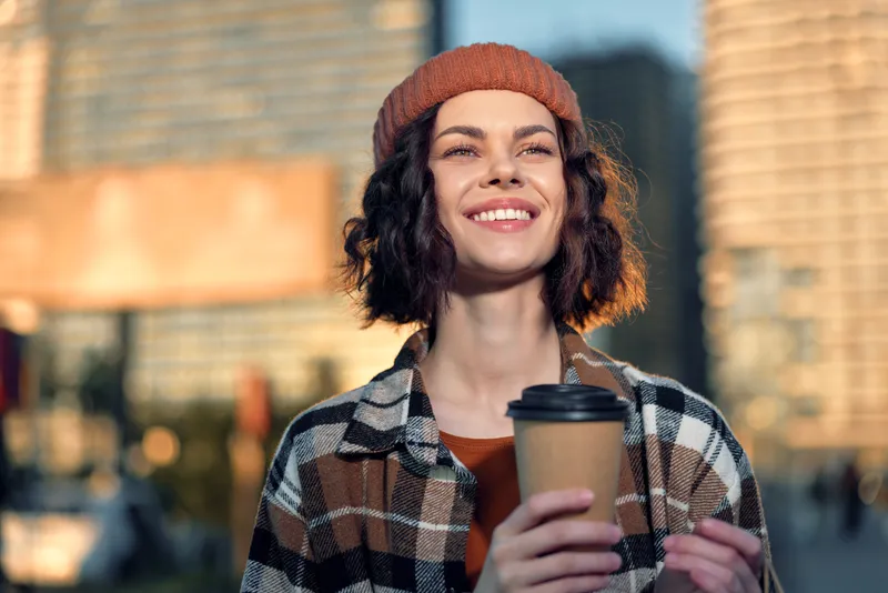 Woman with beanie and warm smile holds coffee on a city street at goldenhour, candid urban portrait evoking authenticity, mindful living and emotional storytelling with cozy autumn vibe