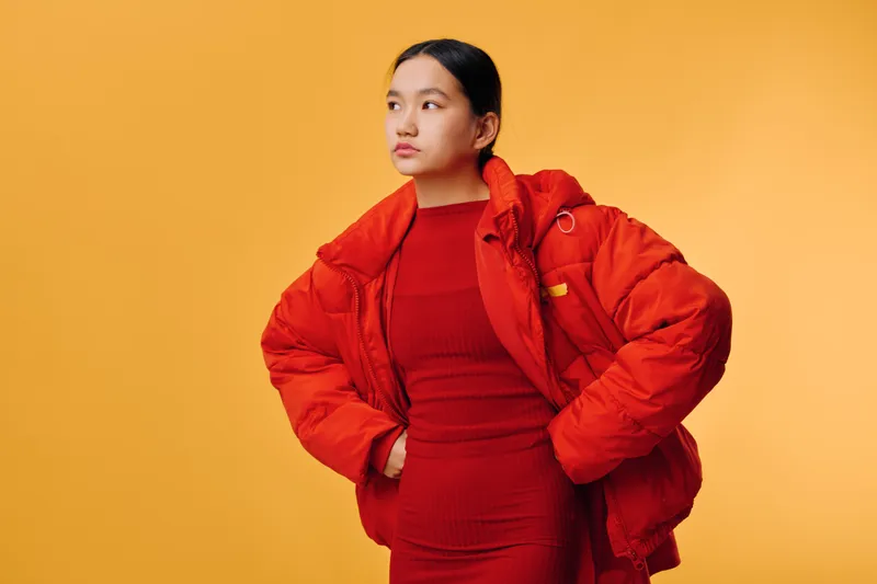 Woman in red dress and oversized jacket poses with hand on hip against bright yellow background. Fashion portrait, casual style, confident look, seasonal outfit for autumn or winter.