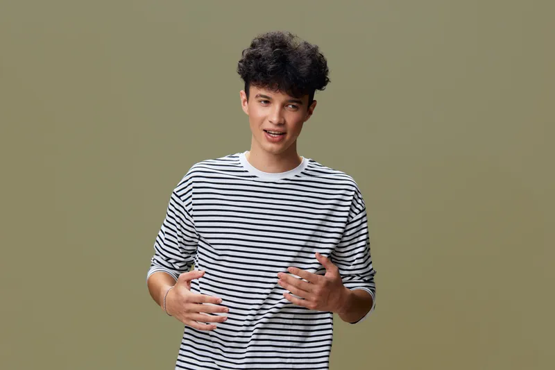 young man in striped shirt expressing emotions with hands, casual style against plain beige background, curly hair, candid moment, natural lighting