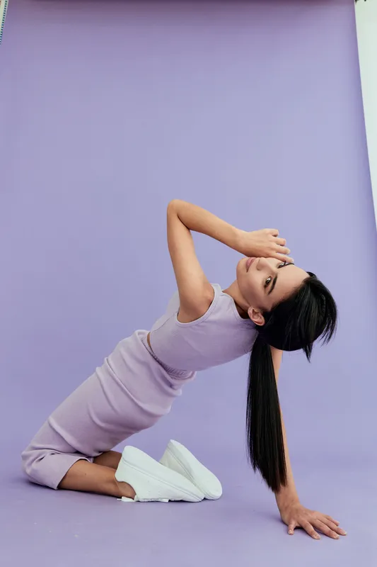 woman posing in lilac outfit kneeling on floor with hand covering face against purple background minimalist fashion style